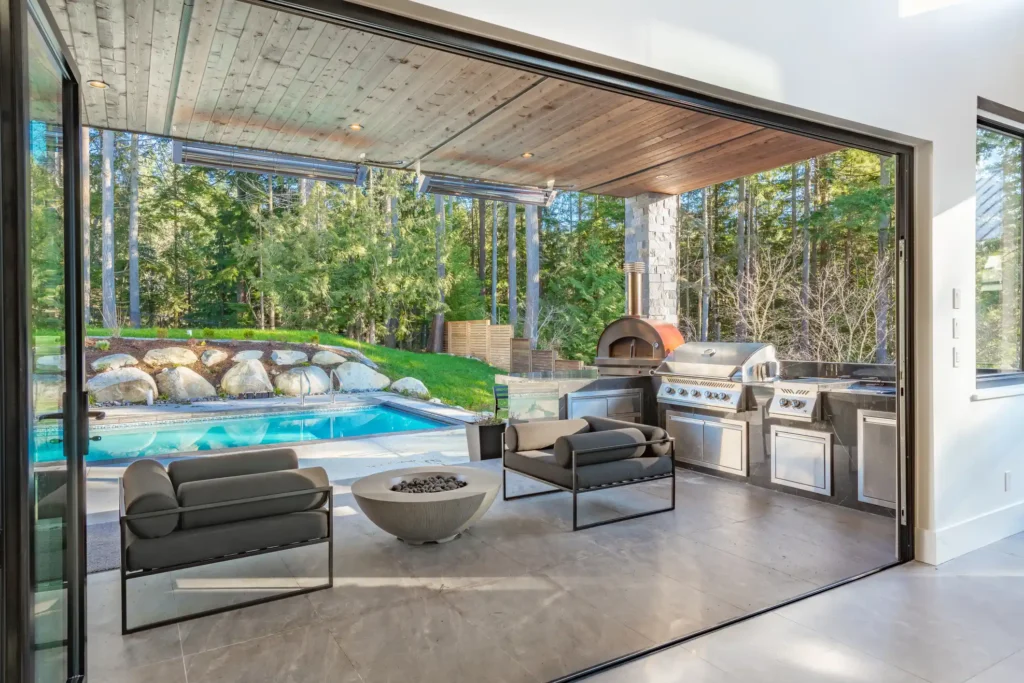 A luxurious covered patio with an outdoor kitchen, lounge seating, and a view of a custom swimming pool and wooded backyard.